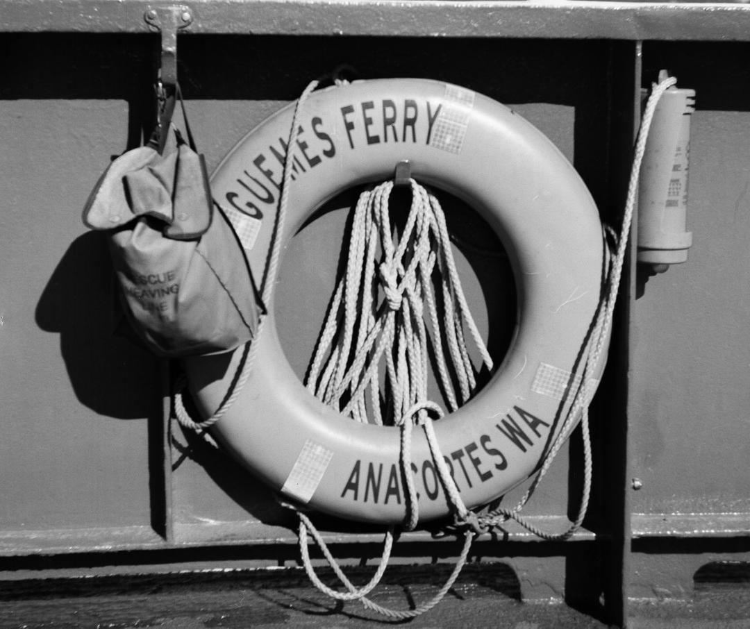 A life ring reading GUEMES FERRY, ANACORTES WA, hanging on the ferry railing with a coiled rescue heaving line, Guemes Channel and the island hills in the background.